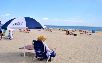 Woman in a straw hat sitting on a chair on the beach next to a beach umbrella. 